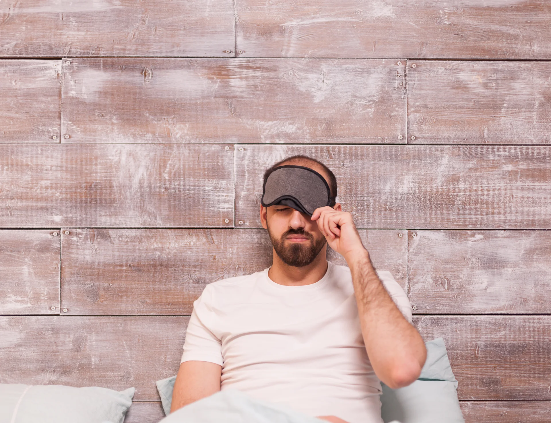 Mann im Bett, lehnt sitzend an eine Wand und hebt seine Schlafmaske halb hoch